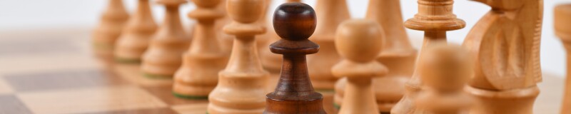 Light coloured wooden chess pieces on a wooden board. Centre pawn is darker coloured wood.