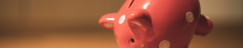 Pink ceramic piggy bank, with white spots. On a wooden floor, balanced on its front legs with its snout on a pile of coins