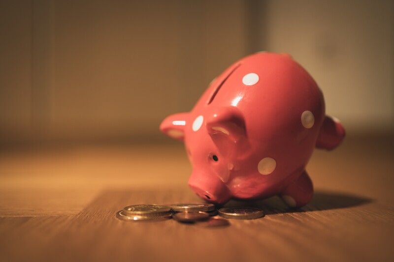 Pink ceramic piggy bank, with white spots. On a wooden floor, balanced on its front legs with its snout on a pile of coins