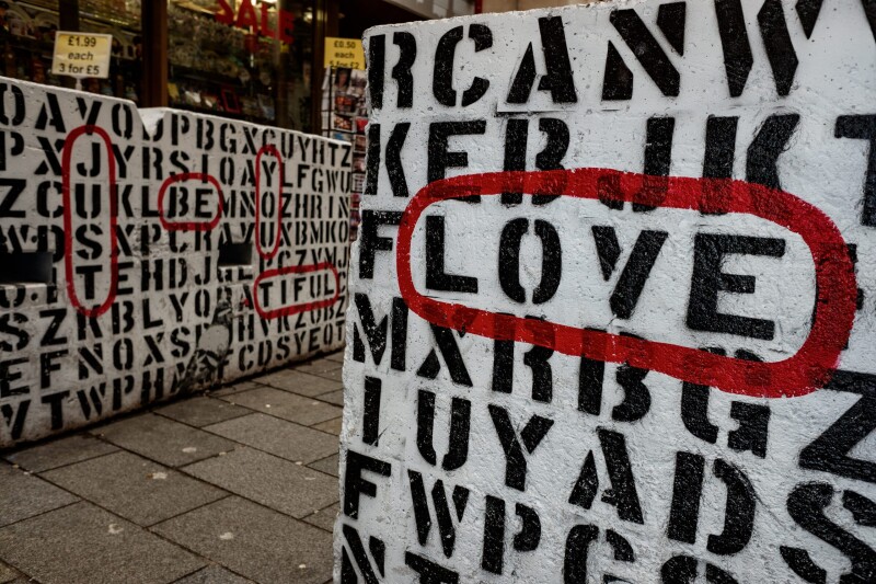 Street art in an alley showing a large-scale black on white word find puzzle, with the words "love" and "just be you tiful" circled in red