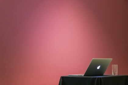 Open Apple laptop and a glass on a table, blush coloured wall behind the table.