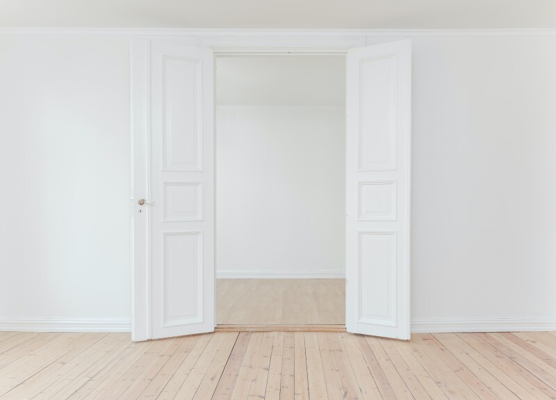 View of an empty room with pale timber flooring, looking through open, white double doors in a white wall leading into another empty room with white walls and pale timber floors