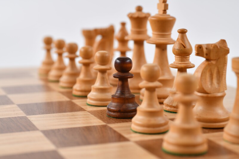 Light coloured wooden chess pieces on a wooden board. Centre pawn is darker coloured wood.