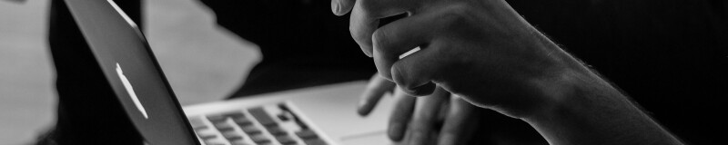 Black and white profile image of the bottom half of a person, seated, with an apple laptop on their lap; their right arm is slightly lifted and in focus, their left hand is on the trackpad