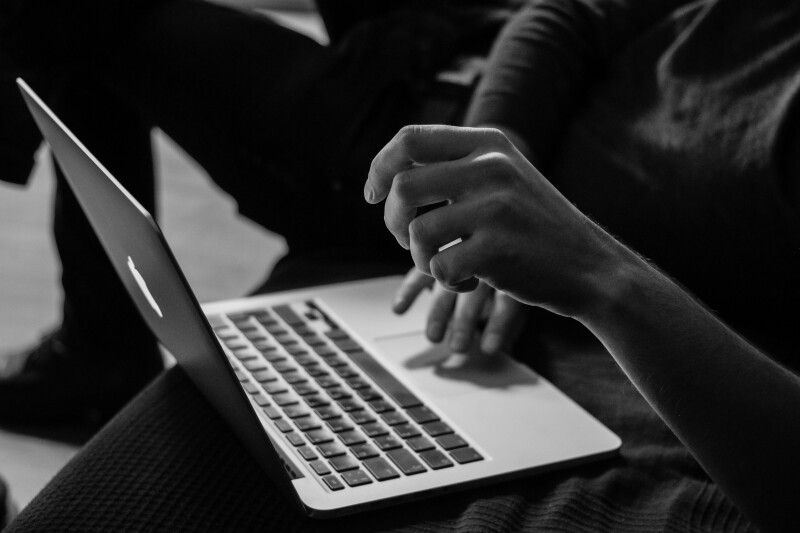 Black and white profile image of the bottom half of a person, seated, with an apple laptop on their lap; their right arm is slightly lifted and in focus, their left hand is on the trackpad