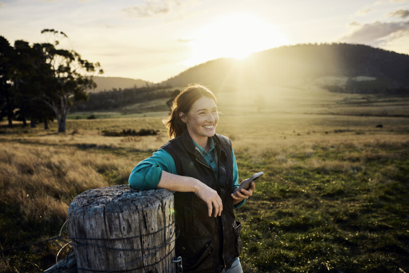 Woman leaning on a fence post in the country holding a device in one hand