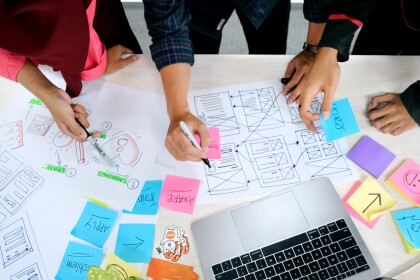 The hands and arms of three people working at a table with UX artefacts, post its and an open laptop
