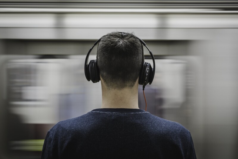 The back view of a person with short hair and headphones on, with a blurred train moving past the front of them