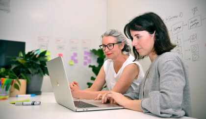 Two people looking at a laptop with post-it notes and drawings on the wall behind.