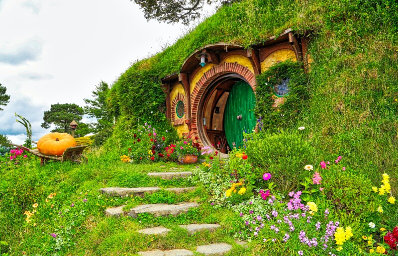 View of Bilbo Baggins' pathway and front door of his hobbit-hole in The Shire. There are pretty wildflowers on the right and left of the path and a giant pumpkin in a wooden wheelbarrow to the left of the green circular front door.