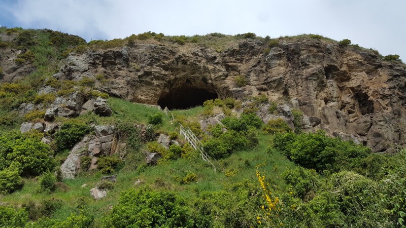 Looking up at rocky cliff face with a cave in it. A handrail, indicating steps, leads up to the cave.