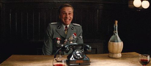Gif of man in military dress grinning broadly and jiggling his shoulders from right to left. He's seated at a table that has an old dial phone on it, a bottle of wine and two glasses with red wine in them