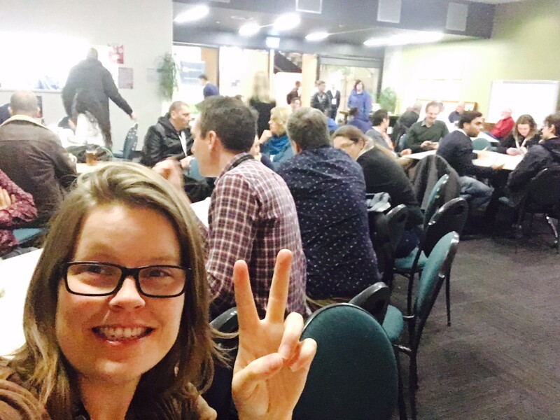 Selfie of woman wearing glasses doing the peace sign, with a room of people sitting at tables behind her