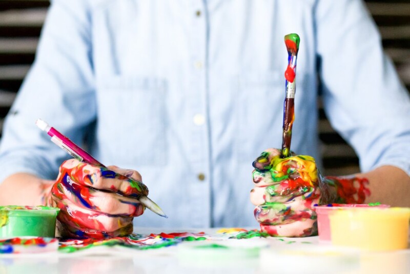 A person sitting at a table holding a pen and a paint brush with paint on their hands, paint brush and table