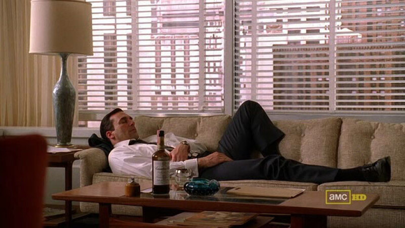 Don Draper from Mad Men asleep on a couch, a bottle of Canadian Club, a glass and an ashtray are on the coffee table.