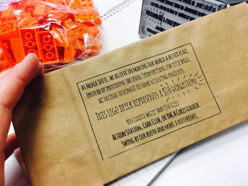 Brown envelope with the text "At Media Suite, we believe in making our works a better place. Instead of providing the usual sponsor swag for these bags, we decided to donate to some deserving charities. This lego brick represents a $10 donation. You choos