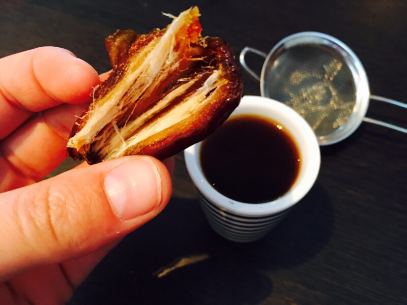 Fingers holding half a fresh date - a small cup of coffee and a strainer on the table