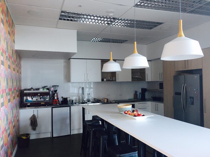 An office kitchen, with long table in the middle, a fruit bowl on it, three lights hanging above it. 