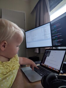 Two screens and a laptop on a table, baby being held and reaching out to press keys on the laptop keyboard