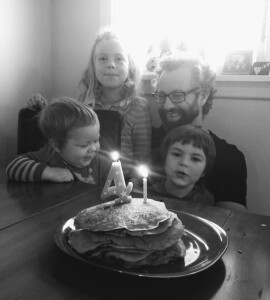 Black and white image of dad crouching holding one child with another child standing to his right, and another child sitting on a chair in front of the standing child. There's a plate of pancakes on the table in front of them. The pancakes have two lit ca