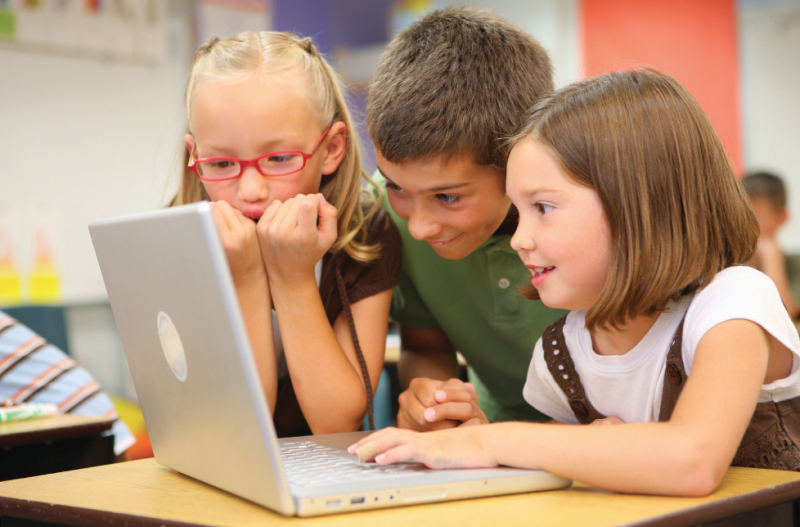 Three primary school aged children huddling around an open laptop on a table