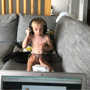 A child with headphones on sitting on a couch. Taken from the perspective of Mum sitting at the other end of the couch, over the top of her laptop.
