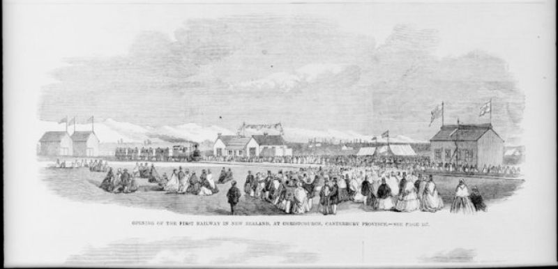 Black and white image with buildings in the background, railway line in the middle ground and people gathered in a field in the foreground.