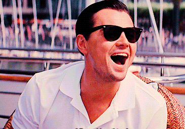 Gif of a man wearing sunglasses, sitting in a cane chair leaning forward and laughing. 