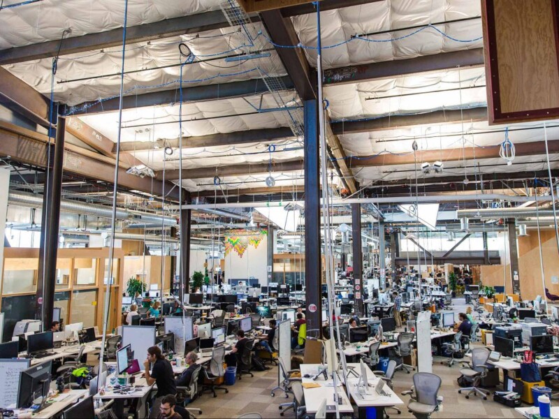 Large, open plan office with an industrial feel, many desks and cables dropping from the ceiling to the desks