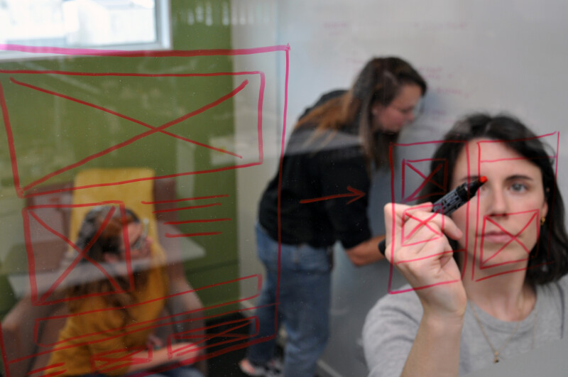 A person writing on glass with a whiteboard marker while two colleagues discuss