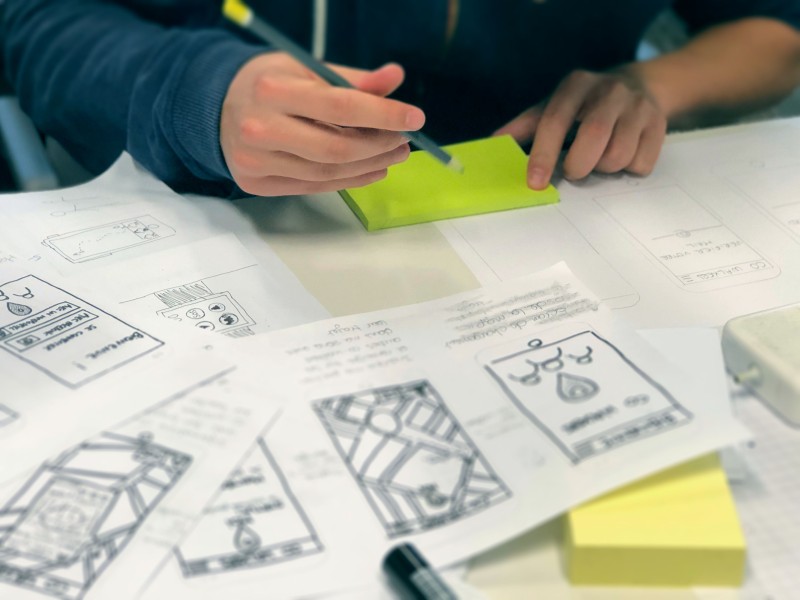 Person writing on a post it note with various hand drawn wireframes of an interface scattered over a desk